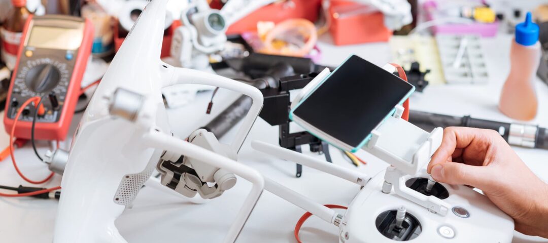 Process is in full swing. Close up of table full of technical equipment and mans hands tuning drone remote controller and landing gears while using smartphone.